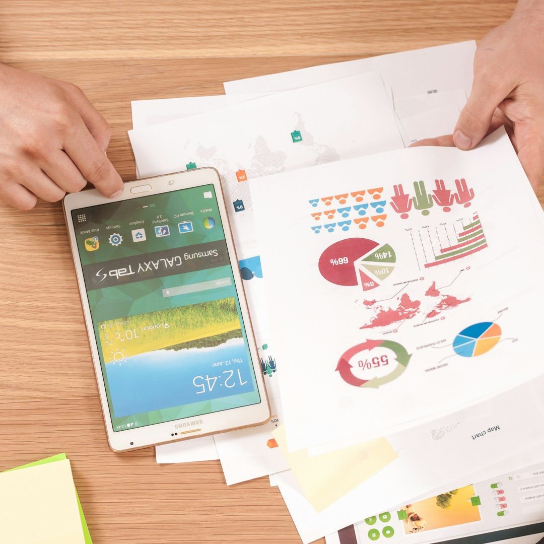 Hände halten ein Tablet neben verschiedenen Grafiken und Notizen -Keywords-Recherche auf einem Tisch.